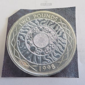 May include: A silver two pound coin with a circular design featuring a stylized sun with a central circle and a date of 1998.