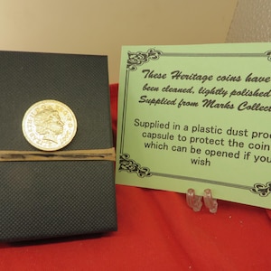 May include: A gold-coloured coin featuring the profile of Queen Elizabeth II and the year 2007, attached to a black textured box with a rubber band. Next to it is a light green card with black text detailing the coin's heritage and storage.