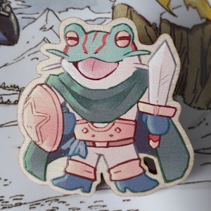 May include: A wooden cutout of a frog wearing armor, holding a sword and shield. The frog is green with red stripes and has a pink tongue sticking out. The armor is brown and the shield is pink with a white star.
