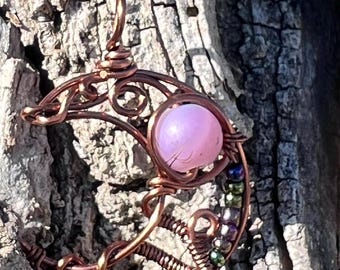 May include: A copper wire pendant in a crescent moon shape, featuring a large pink gemstone at the center. The pendant is adorned with small, colorful beads and intricate wirework, creating a unique, handcrafted design.