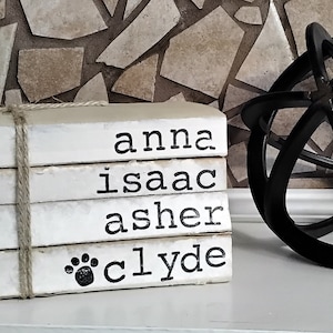 May include: A stack of four white books with the names "anna", "isaac", "asher", and "clyde" printed on them. The books are tied together with twine and a paw print is on the bottom book.