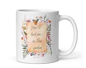 Może przedstawiać: Biały ceramiczny kubek z kwiatowym wzorem i napisem "You'll find me in the garden". Projekt przedstawia ilustrację w stylu akwareli kwiatów i liści w odcieniach różu, zieleni i żółci. Kubek ma zakrzywiony uchwyt.