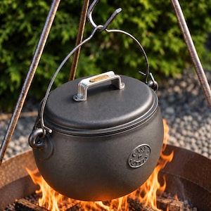May include: A black cast iron cauldron with a matching lid, featuring a metal handle suspended from a tripod. The lid has a silver handle with a small oval label, and the side of the pot displays a circular emblem with the text Cast in Style.