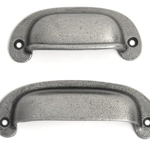 May include: Two antique-style, grey metal drawer pulls. Each pull has a curved, cup-like design with two mounting holes. The metal has a textured, speckled appearance, suggesting an aged or weathered finish. Suitable for cabinets or drawers.