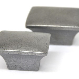 May include: Two square-shaped cabinet knobs made of metal with a brushed nickel finish. The knobs have a slightly raised edge around the top and bottom.