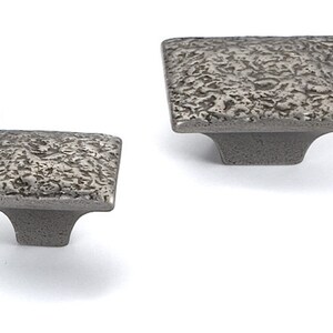 May include: Two square cabinet knobs with a hammered metal finish. The knobs are made of metal and have a square base with a raised square top.