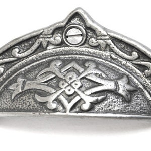 May include: A silver-coloured, ornate drawer pull with a curved, half-moon shape. The decorative hardware features detailed scrollwork and a central design with a cross-like element. Two screws secure the pull to the drawer.