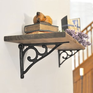 May include: A wooden shelf with decorative black metal brackets is mounted on a white wall. The shelf holds a stack of books, a wooden fox figurine, and a sprig of purple flowers. The shelf is a dark brown colour.