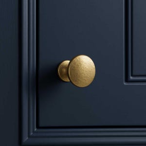 May include: A close-up of a gold cabinet knob with a textured finish. The round knob is mounted on a dark blue cabinet door. The hardware is a simple, yet elegant design.