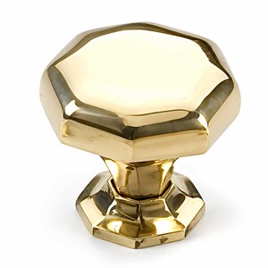 May include: A polished gold-coloured metal cabinet knob featuring a multi-faceted, octagonal top surface and a matching octagonal base. The hardware has a reflective, smooth finish with distinct geometric edges and a tapered, central neck connecting the top.