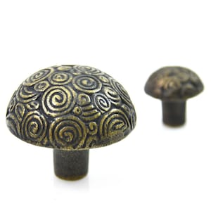 May include: Two antique brass mushroom-shaped cabinet knobs with a swirling pattern. The larger knob is in focus and the smaller knob is out of focus in the background.