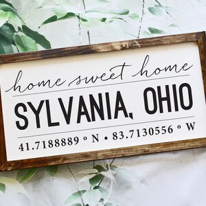 May include: A rectangular wooden sign with a dark brown frame. The sign has a white background with the words "home sweet home" in cursive script. Below, in bold black letters, it reads "SYLVANIA, OHIO" with the coordinates 41.7188889° N • 83.7130556° W.