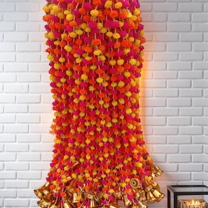 May include: A long garland of colorful pom-poms in pink, yellow, and orange, with gold bells hanging from the bottom. The garland is hanging on a white brick wall.