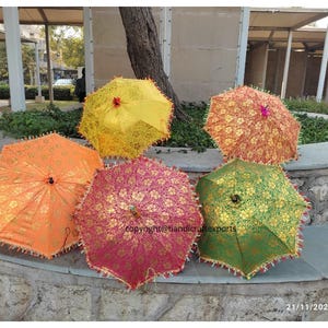 May include: Five colourful patterned umbrellas with fringe trim. The umbrellas are arranged in a semi-circle on a stone surface. The umbrellas are yellow, orange, pink, green, and blue.