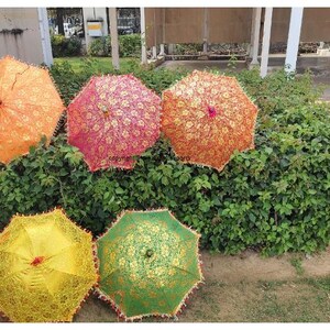 May include: Five colourful lace umbrellas with intricate patterns. The umbrellas are orange, pink, yellow, and green. They are arranged in a semi-circle on a bed of green foliage.
