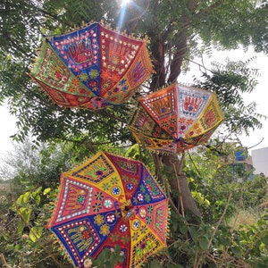 Op de afbeelding: Drie kleurrijke, gepatroneerde parasols die aan een tak hangen. De parasols zijn versierd met handgestikte ontwerpen, waaronder bloemen, geometrische vormen en spiegelachtige versieringen.