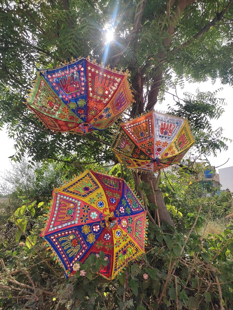 Op de afbeelding: Drie kleurrijke, gepatroneerde parasols die aan een tak hangen. De parasols zijn versierd met handgestikte ontwerpen, waaronder bloemen, geometrische vormen en spiegelachtige versieringen.