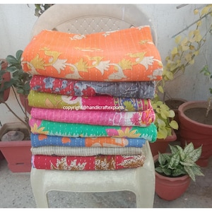 Può includere: Una pila di otto quilt kantha colorati, realizzati con sari riciclati, è esposta su una sedia bianca. I quilt sono sovrapposti in una varietà di motivi e colori, tra cui arancione, rosa, verde, blu e rosso.