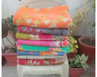May include: A stack of eight colorful kantha quilts, made from recycled saris, are displayed on a white chair. The quilts are layered in a variety of patterns and colors, including orange, pink, green, blue, and red.