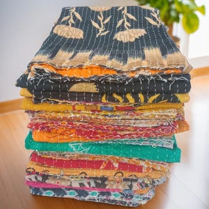 May include: A stack of colorful kantha quilts, featuring a variety of floral and geometric patterns. The quilts are made of recycled saris and are hand-stitched with kantha embroidery.