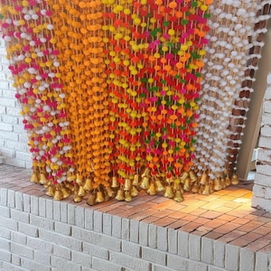 May include: A colorful garland of pom-poms with gold bells hanging from the bottom. The garland is made of white, yellow, orange, pink, and green pom-poms.