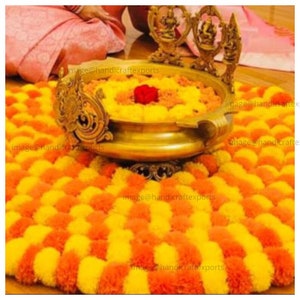 May include: A traditional Indian wedding ceremony decoration with a brass bowl filled with yellow and orange marigold flower petals. The bowl is surrounded by a circular arrangement of marigold flower pom-poms.