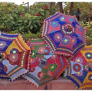 May include: Five colourful, patterned umbrellas with fringe trim. The umbrellas are made of fabric and have a variety of colours and designs, including blue, red, yellow, green, and white. The umbrellas are arranged in a row on a pink surface.
