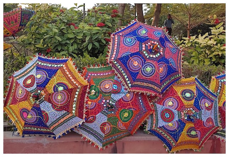 May include: Five colourful, patterned umbrellas with fringe trim. The umbrellas are made of fabric and have a variety of colours and designs, including blue, red, yellow, green, and white. The umbrellas are arranged in a row on a pink surface.