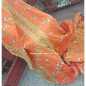 以下が含まれることがあります： オレンジ色のカンタキルトで、花柄と白と黄色のアクセントが付いています。キルトは折り畳んでプランターの上にかけられています。