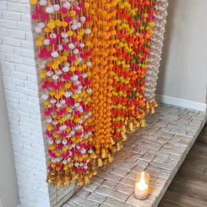 May include: A white brick fireplace decorated with colorful pom-pom garlands with gold bells. The garlands are in shades of pink, yellow, orange, and green.