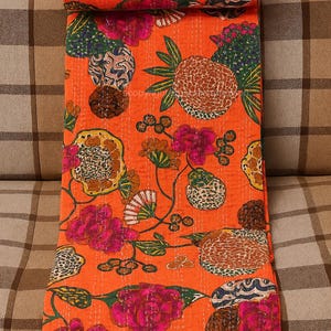 May include: An orange floral quilt with a repeating pattern of pink flowers, brown pineapples, and green leaves. The quilt is folded and placed on a brown and tan plaid couch. The quilt is made of a soft, quilted material.
