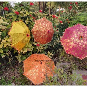 Op de afbeelding: Drie decoratieve parasols met gouden en bloemmotieven. De parasols zijn geel, oranje en roze. Ze worden getoond tegen een achtergrond van groen gebladerte.