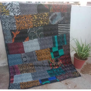 May include: A patchwork quilt with a variety of patterned fabric squares in shades of black, blue, red, green, and gold. The quilt is displayed outdoors, with a small potted plant in the background. The quilt has a stitched design.
