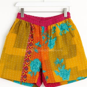 Può includere: Pantaloncini patchwork colorati con fascia in vita rosa e stampa floreale. I pantaloncini presentano un mix di tessuti gialli, arancioni e rosa con motivi floreali blu e cuciture bianche. Appesi a una gruccia bianca.