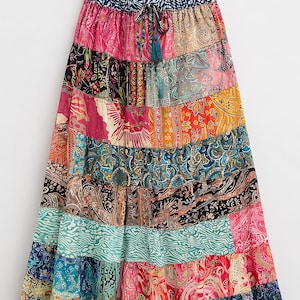 May include: A long, tiered skirt with a patchwork design. The skirt features a variety of colorful patterns and prints, including paisley, floral, and geometric designs. The skirt has a drawstring waist and a tassel detail. The skirt is hanging on a wooden hanger.