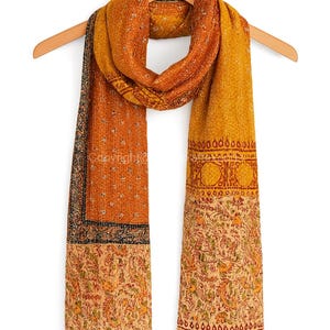 May include: A long, rectangular scarf with a warm color palette of orange, yellow, and beige. The scarf features intricate patterns and designs, including floral and geometric motifs. It is displayed on a wooden hanger.