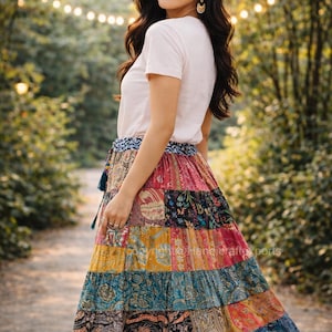 May include: A woman wearing a white t-shirt and a long, patchwork skirt with a variety of colors and patterns. The skirt has a tiered design and a blue waistband. The background is a blurred outdoor scene with trees and soft lighting.