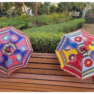 Op de afbeelding: Twee kleurrijke, gepatroneerde parasols met franjes. De parasols zijn gemaakt van stof en hebben een verscheidenheid aan kleuren, waaronder rood, blauw, geel en groen. Ze zijn versierd met pailletten en spiegels. De parasols staan op een houten bank.