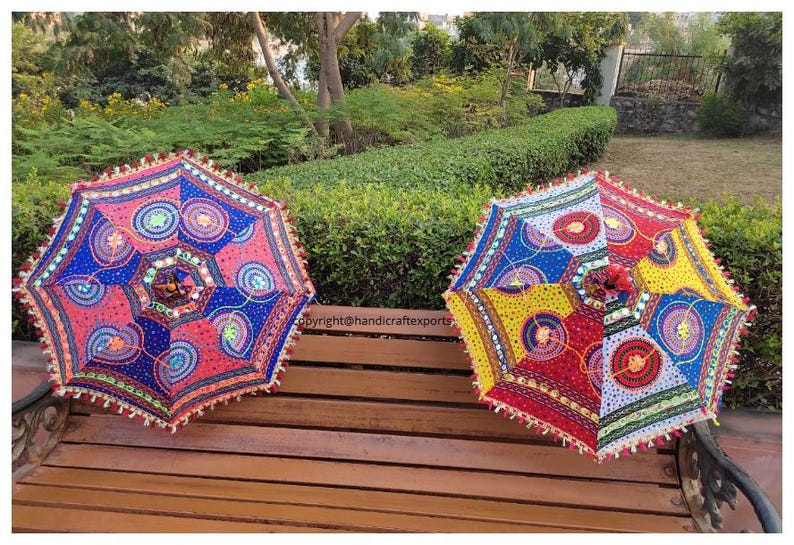 Op de afbeelding: Twee kleurrijke, gepatroneerde parasols met franjes. De parasols zijn gemaakt van stof en hebben een verscheidenheid aan kleuren, waaronder rood, blauw, geel en groen. Ze zijn versierd met pailletten en spiegels. De parasols staan op een houten bank.