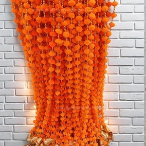 Puede incluir: Guirnalda de flores de caléndula naranja con cascabeles dorados colgando de una pared de ladrillo blanco. La guirnalda está hecha de pompones y cuentas.