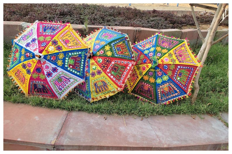 May include: Three colourful umbrellas with embroidered designs, including elephants, flowers, and geometric patterns. The umbrellas are made of fabric and have a fringe trim.