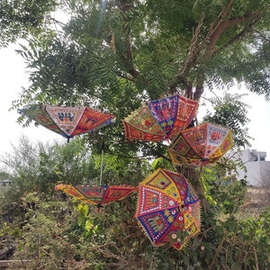 Op de afbeelding: Vijf kleurrijke, gepatroneerde paraplu's hangen aan een tak van een boom. De paraplu's zijn gemaakt van stof en hebben ingewikkelde borduursels en versieringen. Ze zijn een levendige mix van rood, geel, blauw, groen en roze.