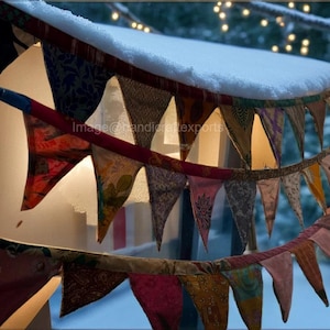 Puede incluir: Guirnalda de tela colorida con banderines triangulares en varios patrones y colores, ensartados en un cordón rojo. La guirnalda está colgada sobre un alféizar cubierto de nieve, con un fondo invernal borroso.