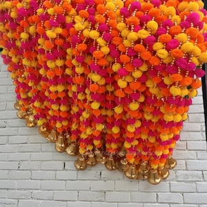 Puede incluir: Una guirnalda de pompones coloridos en amarillo, naranja y rosa, con campanillas doradas colgando de la parte inferior. La guirnalda está colgada en una pared de ladrillo blanco.