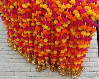 May include: A garland of colorful pom-poms in yellow, orange, and pink, with gold bells hanging from the bottom. The garland is hanging on a white brick wall.
