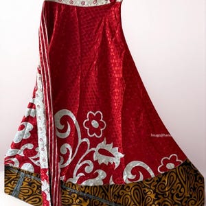May include: A vibrant red skirt with a white floral and swirl pattern. The skirt has a patterned border in shades of gold and black. The top of the skirt has a white and red patterned design. The skirt is hanging on a hanger.