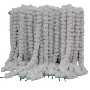 May include: White artificial flower garland made of pom poms. The garland is hanging in a vertical line and is made of many small white pom poms.