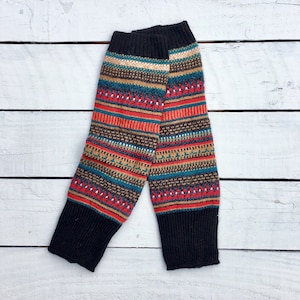 May include: Knitted leg warmers with a black cuff and a colourful pattern. The pattern includes red, teal, tan, and white stripes and geometric designs. The leg warmers are displayed on a white wooden surface.
