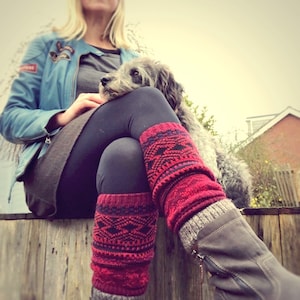 Può includere: Una donna indossa una giacca di jeans blu e leggings neri con scaldagambe a motivi rossi e neri. È seduta su una superficie di legno con un piccolo cane grigio che riposa sulle sue ginocchia.
