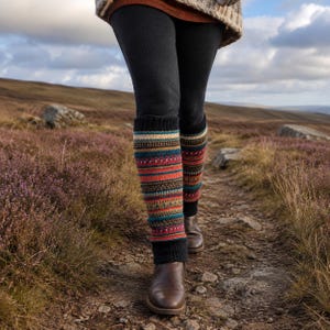 Puede incluir: Un par de calentadores de punto con un patrón de rayas horizontales multicolor en tonos rojo, azul, amarillo y marrón, con puños acanalados negros sólidos en la parte superior e inferior, usados sobre leggings negros y botas de cuero marrón.